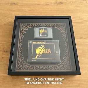 Könnte beinhalten: Gerahmtes Nintendo 64 Spielmodul und Box-Set. Das Modul ist grau mit einem goldenen Logo. Die Box hat ein goldenes und schwarzes Design mit dem Spieltitel. Der Rahmen ist schwarz mit einem dekorativen goldenen Rand.