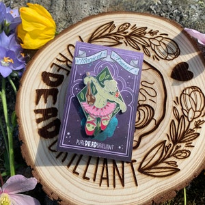 May include: A green and pink enamel pin featuring a whimsical creature wearing a tutu and holding a bottle. The pin is on a purple card with the text "Magical Enamel" and "Pure Dead Brilliant".