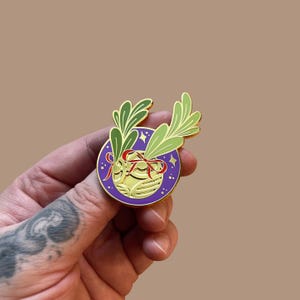 Könnte beinhalten: Goldene Emaille-Anstecknadel mit einem violetten Kreis, in dem sich eine goldene Kugel befindet. Die Kugel ist mit einer roten Schleife und grünen Blättern verziert. Der Hintergrund ist ein violetter Kreis mit weißen Sternen.