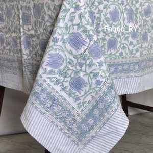 May include: White tablecloth with a blue floral pattern and a striped border. The floral design features blue tulip-like flowers and green leaves. The border has a repeating floral pattern and blue and white stripes.
