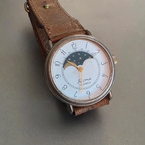May include: A vintage wristwatch with a white face, gold hands, and a moon phase indicator. The watch has a silver case and a brown leather strap. The watch face displays the brand name "Fima" and "17 Jewels".