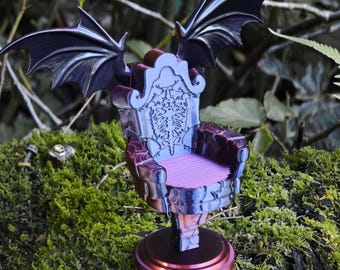 May include: A miniature throne with bat wings, in shades of purple and black, sits on a mossy surface. The chair features a detailed backrest and a pink seat. The wings are spread out, adding to the gothic aesthetic.