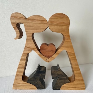 May include: Wooden sculpture featuring two figures holding a heart, with two cat figures at the base. The figures are made of light brown wood, with a darker brown heart and cat figures. A unique home decor item.