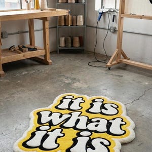 Hand-tufted doormat with bright yellow “It Is What It Is” lettering on a neutral background, rectangular design with textured fibers; stylish and durable for home entryways, hallways, or living spaces, perfect for adding personality to decor.