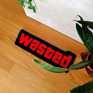 Op de afbeelding: Een rechthoekig zwart tapijt met het woord "wasted" in rode, vette letters. Het tapijt ligt op een houten vloer, met groene planten op de achtergrond.