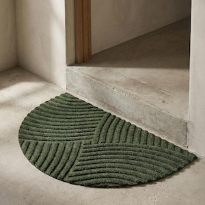 May include: An olive green, semi-circular doormat with a textured geometric design. The mat is positioned in front of a doorway, with a concrete floor and steps. The mat's pattern consists of curved lines.