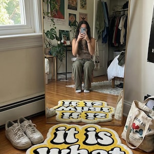 Hand-tufted rectangular doormat with bright yellow “It Is What It Is” lettering, featuring a soft, textured pile and sturdy backing; ideal for welcoming guests while adding a bold, humorous statement to home decor.