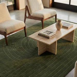 May include: A modern living room scene featuring two cream-colored armchairs with wooden frames, a light-colored rectangular coffee table, and a green patterned rug. A small vase and two books are on the table. Natural light streams in from a large window.