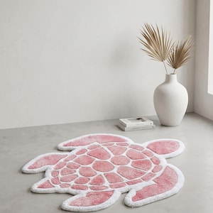 Puede incluir: Una alfombra de baño rosa y blanca con forma de tortuga. La alfombra presenta un patrón texturizado en forma de guijarros rosas, delineado con un borde blanco. Está colocada sobre un suelo gris claro en una habitación con una pared blanca y un jarrón.