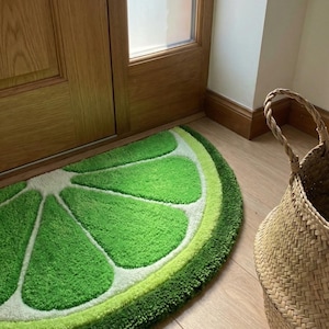 Può includere: Un tappeto a forma di fetta di lime, verde brillante e con superficie strutturata. Il tappeto presenta un centro bianco e una buccia verde chiaro, creando un design realistico di frutta. Vicino a una porta di legno e un cesto di vimini.
