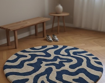 May include: A round rug with a blue and beige abstract pattern. The rug is centered on a wooden floor, with a wooden bench and small table in the background. The rug's design features flowing, organic shapes.