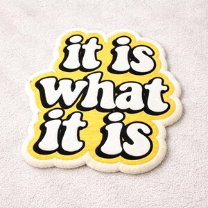 Soft tufted doormat featuring the bold phrase “It Is What It Is” in yellow glow letters, rectangular shape, and sturdy construction; adds a playful, motivational touch to home interiors while remaining functional and durable.