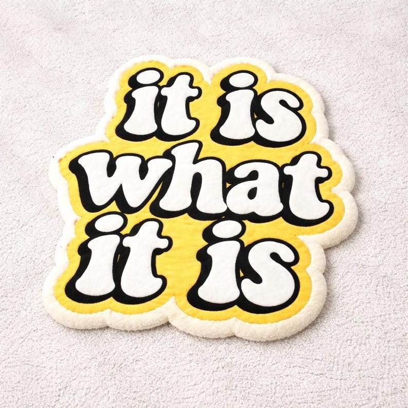 Soft tufted doormat featuring the bold phrase “It Is What It Is” in yellow glow letters, rectangular shape, and sturdy construction; adds a playful, motivational touch to home interiors while remaining functional and durable.