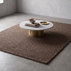 May include: A round coffee table with a white marble top and a cylindrical brass base. On the table sits a small beige bowl, two stacked books, and a small gold tray. The table is placed on a large, rectangular, brown textured wool rug.