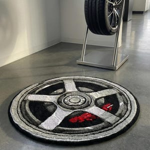 Puede incluir: Una alfombra redonda con un diseño de rueda de coche en tonos grises, negros y rojos. Un neumático de tamaño completo se exhibe en un soporte metálico detrás de la alfombra. La alfombra presenta una representación detallada de una rueda de coche.
