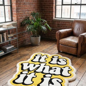 May include: A yellow rug with the phrase "it is what it is" in white and black lettering. The rug has a cloud-like shape and is placed on a wooden floor. The room has exposed brick walls and large windows.