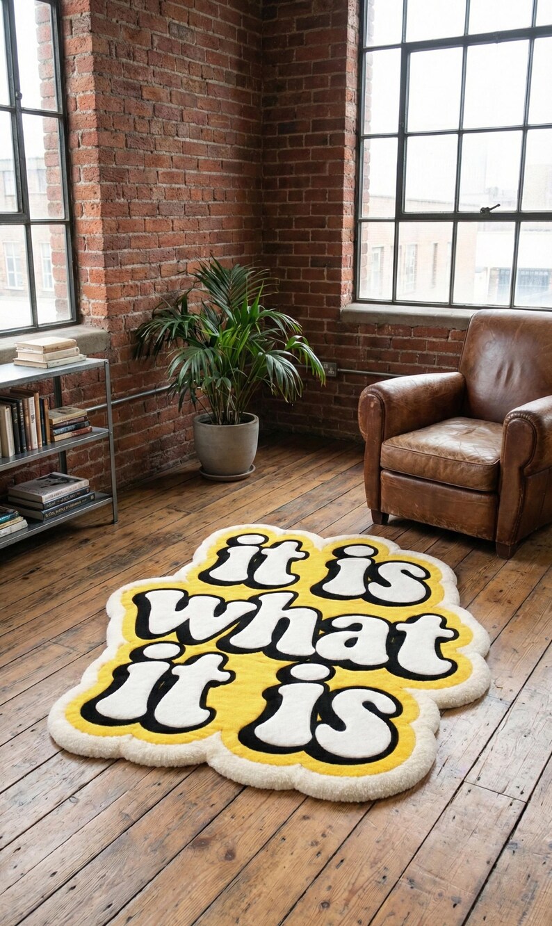 May include: A yellow rug with the phrase "it is what it is" in white and black lettering. The rug has a cloud-like shape and is placed on a wooden floor. The room has exposed brick walls and large windows.