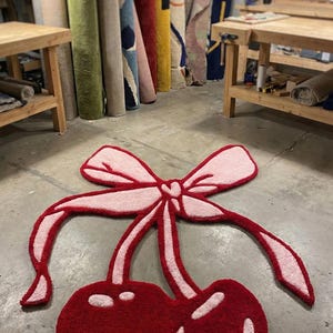 Può includere: Un tappeto rosso a forma di ciliegia con un fiocco rosa. Il tappeto è realizzato in un materiale morbido e strutturato e presenta accenti bianchi sulle ciliegie. Sullo sfondo sono visibili tappeti arrotolati.