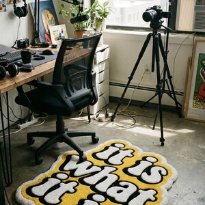 Close-up of a custom tufted doormat with glowing yellow “It Is What It Is” text, showing detailed fibers and plush texture; perfect for adding a stylish and durable accent to entryways, hallways, or living areas.