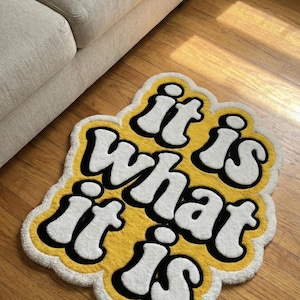 Close-up of a hand-tufted doormat with bold yellow “It Is What It Is” text, showing textured fibers and a sturdy surface; perfect for adding a fun, motivational accent to doors, hallways, or indoor home decor.