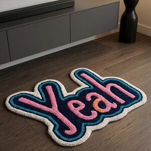 Könnte beinhalten: Dekorativer Teppich mit dem Wort "Yeah" in einem Bubble-Letter-Design. Der Teppich hat einen weißen Rand, eine türkisfarbene Umrandung und rosa Schrift mit einem orangefarbenen Akzent. Er liegt auf einem Holzboden.