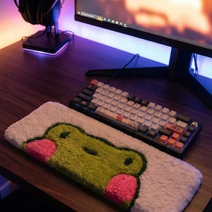 Puede incluir: Una alfombra rectangular blanca con una cara de rana verde y mejillas rosas, colocada sobre un escritorio de madera. Un teclado blanco y negro está al lado de la alfombra. Unos auriculares negros están en un soporte y un monitor de ordenador está en el fondo.