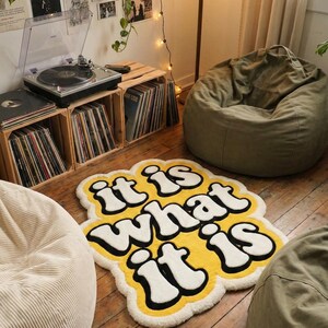 Stylish custom text doormat with “It Is What It Is” in glowing yellow letters on a soft tufted background; rectangular shape fits entryways, living spaces, or bedrooms, combining durability with playful home decor charm.