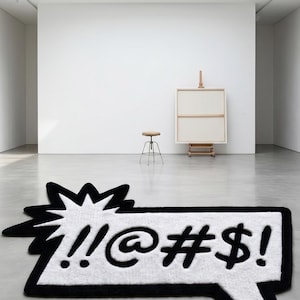 May include: A white and black speech bubble rug with exclamation marks, an at symbol, a hashtag, and a dollar sign. The rug is in a modern art gallery setting with a stool, easel, and a large white wall.
