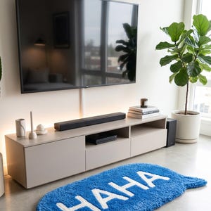 May include: A blue rug in the shape of a speech bubble with the word "HAHA" in white. The rug is on a concrete floor in front of a light-colored entertainment center with a large TV above it. A potted plant is to the right.