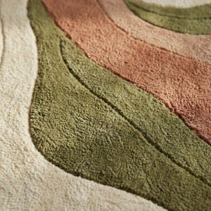 May include: Close-up of a textured rug with a wavy design. The rug features stripes of beige, olive green, and muted coral colors. The soft, plush texture is visible, suggesting a comfortable floor covering.
