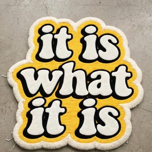 Soft and durable tufted mat displaying the phrase “It Is What It Is” in yellow glow lettering, designed for indoor use in entryways or living rooms; the textured surface provides comfort while enhancing a modern, personalized decor style.