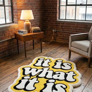 May include: A yellow rug with the phrase "it is what it is" in white, outlined in black. The rug is on a wooden floor in a room with brick walls and large windows. A lamp and side table are visible.
