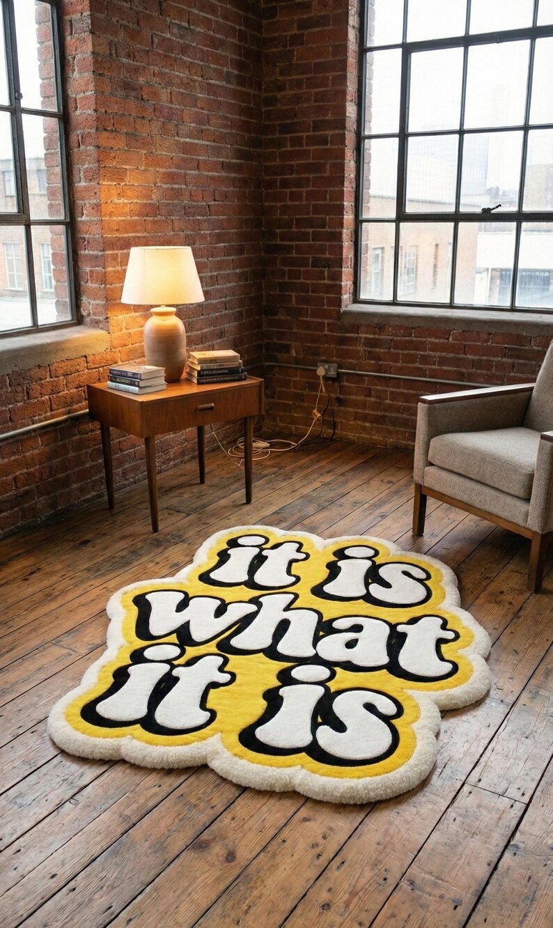 May include: A yellow rug with the phrase "it is what it is" in white, outlined in black. The rug is on a wooden floor in a room with brick walls and large windows. A lamp and side table are visible.