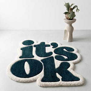 Könnte beinhalten: Ein teal- und cremefarbener Teppich mit den Worten "it's ok" in einer verspielten, abgerundeten Schriftart. Der Teppich hat ein weiches, strukturiertes Aussehen und liegt auf einer weißen Oberfläche. Eine dekorative Pflanze auf einem Sockel befindet sich im Hintergrund.