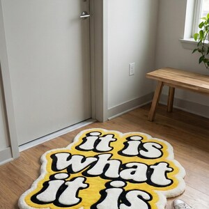 Personalized yellow-lettered “It Is What It Is” tufted mat with a textured, plush surface and rectangular shape; suitable for indoor entryways, living rooms, or decorative floor accents that combine style with function.