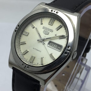 May include: A vintage Seiko 5 automatic watch with a silver-tone case and a cream-colored dial. The watch features rectangular hour markers, a day-date display, and a black leather strap. The watch face displays the text "SEIKO 5" and "AUTOMATIC".