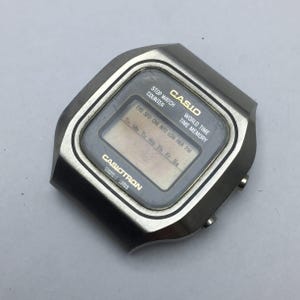 Puede incluir: Un reloj digital Casio Casitron vintage con una caja plateada. La esfera del reloj muestra la hora y el día de la semana. El reloj tiene el texto "CASIO" y "CASIOTRON" impreso en la esfera.