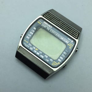 May include: A vintage Seiko digital watch with a silver case and a rectangular display. The watch face features text including "SEIKO", "HOME TIME ALARM", and "WORLD TIME ALARM". The watch has a black band and several buttons.