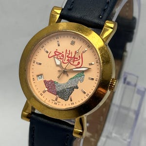 May include: Gold-tone wristwatch with a peach-colored face featuring Arabic script and a map design. The watch has a date display at the 9 o'clock position, a diamond at the 12 o'clock position, and a navy blue leather strap.