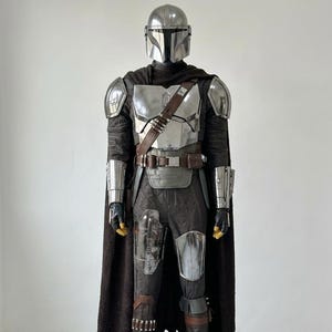 May include: A full-body Mandalorian costume featuring a metallic silver helmet, chest plate, shoulder pauldrons, and gauntlets. The outfit includes a dark brown textured cape, a utility belt with pouches, dark trousers, and brown leather boots.