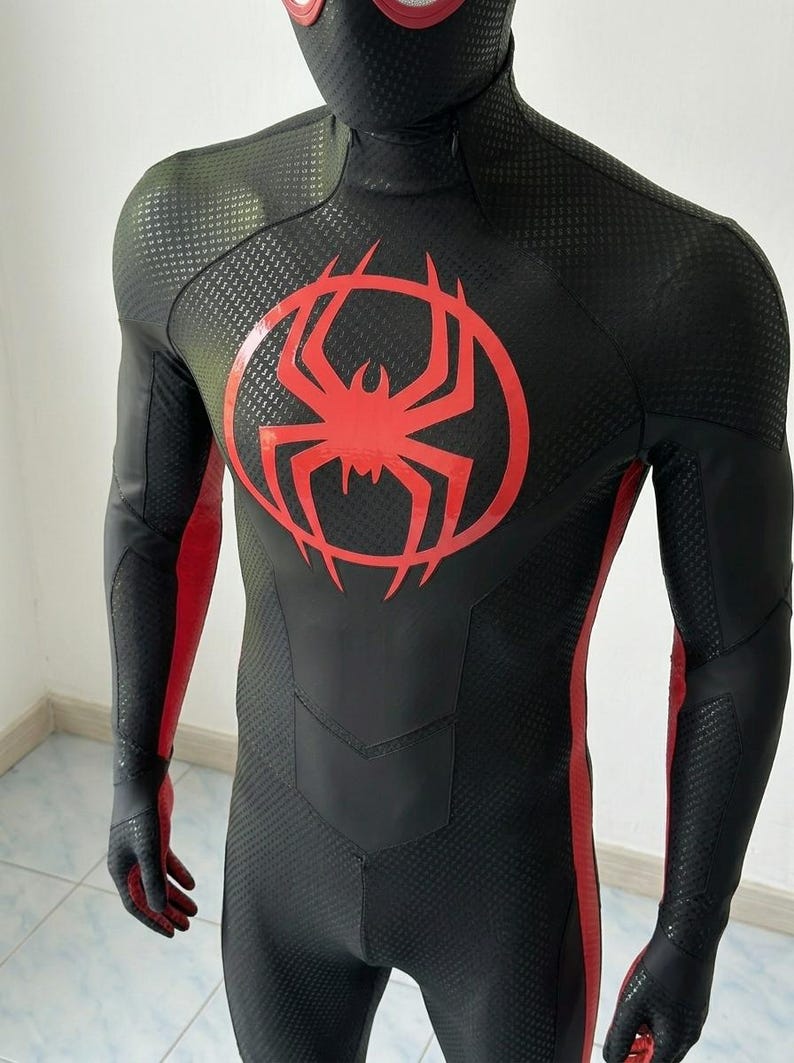 May include: A black full-body suit featuring a textured honeycomb pattern, a prominent red spider emblem on the chest, and red vertical stripes running along the sides of the torso and arms. The suit includes a matching mask with red eye lenses.
