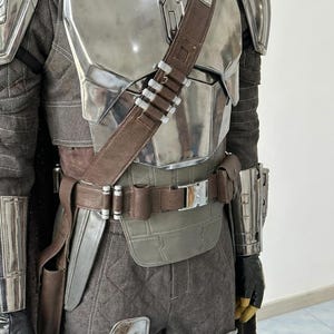 May include: A full-body suit of armor featuring a polished silver chest plate, shoulder pauldrons, and gauntlets. The ensemble includes a brown leather utility belt with a silver buckle, a matching diagonal bandolier, and dark gray quilted trousers.