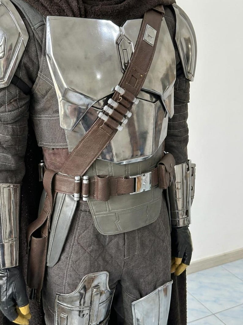 May include: A full-body suit of armor featuring a polished silver chest plate, shoulder pauldrons, and gauntlets. The ensemble includes a brown leather utility belt with a silver buckle, a matching diagonal bandolier, and dark gray quilted trousers.