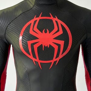 May include: A black and dark olive green long-sleeve bodysuit featuring a high neck with a zipper closure. The chest displays a large, glossy red spider emblem centered within a circular outline. The fabric has a textured, repeating pattern.
