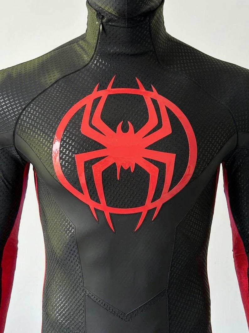 May include: A black and dark olive green long-sleeve bodysuit featuring a high neck with a zipper closure. The chest displays a large, glossy red spider emblem centered within a circular outline. The fabric has a textured, repeating pattern.