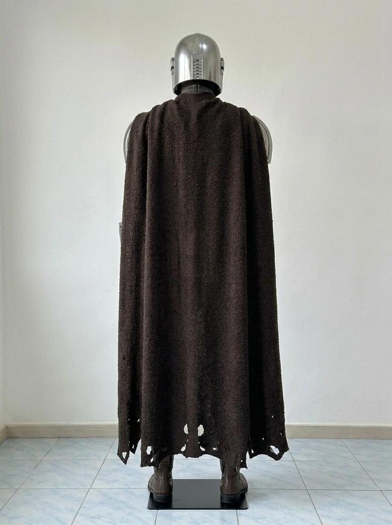 May include: A full-length dark brown textured cape worn over a metallic silver helmet and armor set. The cape features a distressed, frayed hemline with visible holes and uneven edges, draped over the shoulders and extending down to the boots.