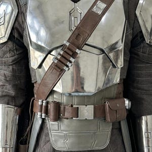 May include: A suit of armor featuring a polished silver chest plate, shoulder pauldrons, and forearm guards. A brown leather bandolier with silver cartridges crosses the chest, paired with a matching brown utility belt and a dark brown quilted tunic.