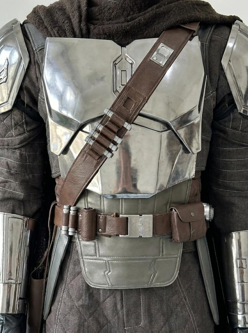 May include: A suit of armor featuring a polished silver chest plate, shoulder pauldrons, and forearm guards. A brown leather bandolier with silver cartridges crosses the chest, paired with a matching brown utility belt and a dark brown quilted tunic.