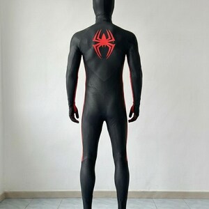 May include: A full-body black spandex suit featuring a textured honeycomb pattern throughout. The back displays a large, stylized red spider emblem centered between the shoulder blades, with red vertical stripes running down the sides of the legs.