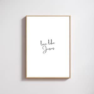 May include: A framed print with the words "love like Jesus" in elegant black script on a white background. The frame is a light wood color, and the print is displayed against a white wall.